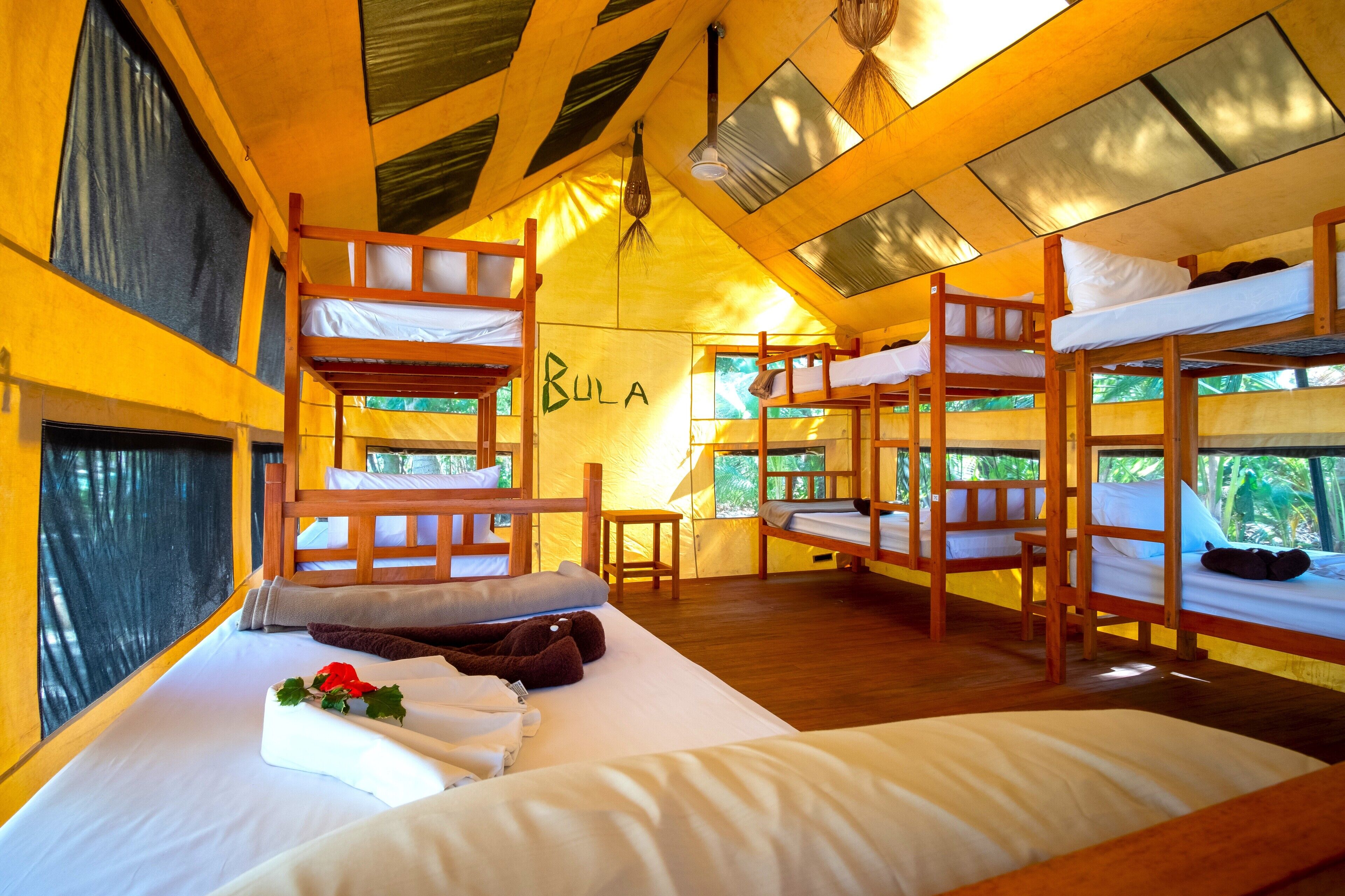 Une chambre de type dortoir avec des lits superposés, une petite table et un mur sur lequel est écrit le mot « BULA ».