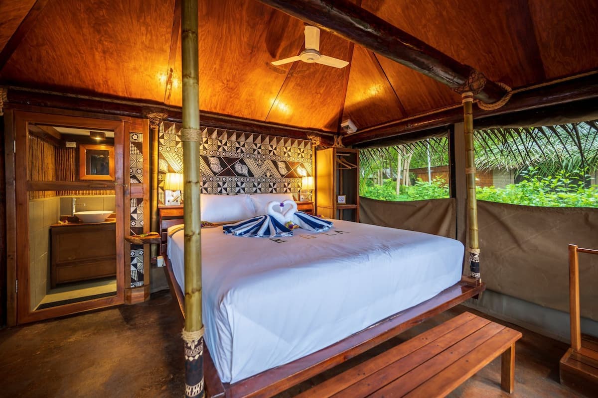 A wooden four-poster bed with a white bedspread and two swan-shaped towels, a wooden bench, a small bedside table with a lamp, and a view of greenery through a bamboo screen.