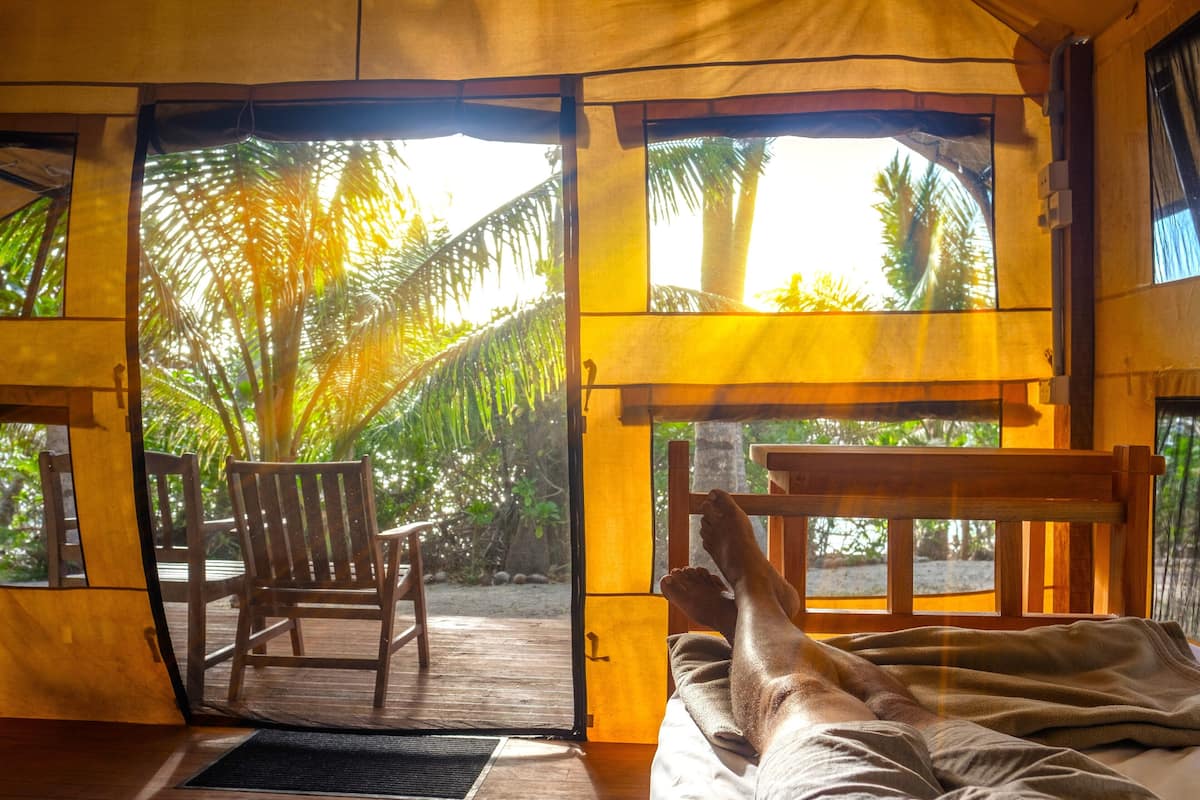 A room with a bed, a chair, and a view of palm trees through open windows.