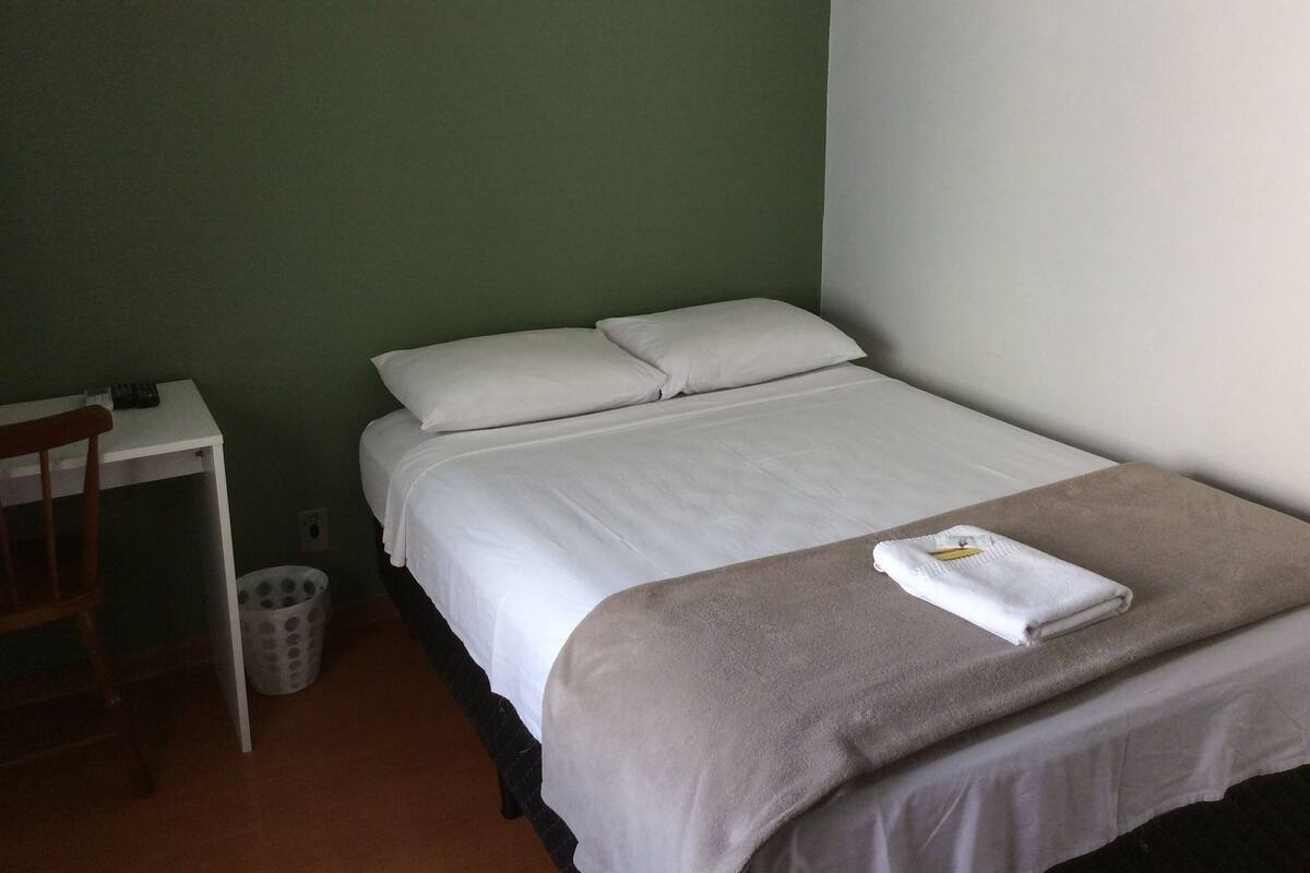 Uma cama de solteiro com roupa de cama branca e um cobertor bege, uma escrivaninha pequena com uma cadeira, e uma lixeira.