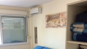A room with two beds, each with a blue patterned bedspread and white pillows. There is a window with blinds, an air conditioning unit on the wall, and a framed picture above the beds. - Pousada Terra Sul (Porto Alegre)