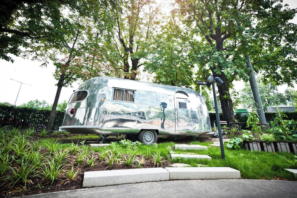 A vintage camper parked in a lush garden with a metal fence and a street lamp nearby.