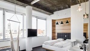 A modern hotel room with a large window, a hanging hammock, a bed with white bedding, a wooden accent wall, and a built-in sink. - Hotel Daniel Vienna (Vienna)