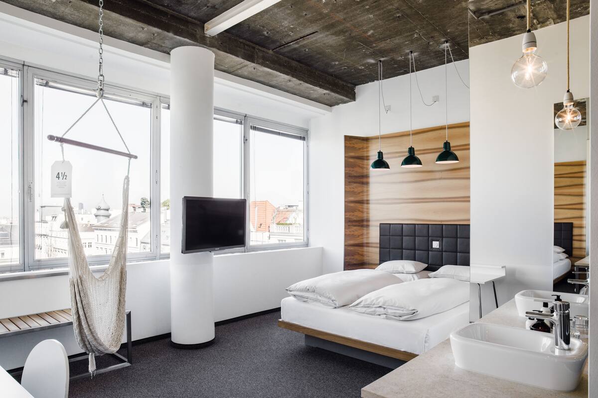 A modern hotel room with a large window, a hanging hammock, a bed with white bedding, a wooden accent wall, and a built-in sink.