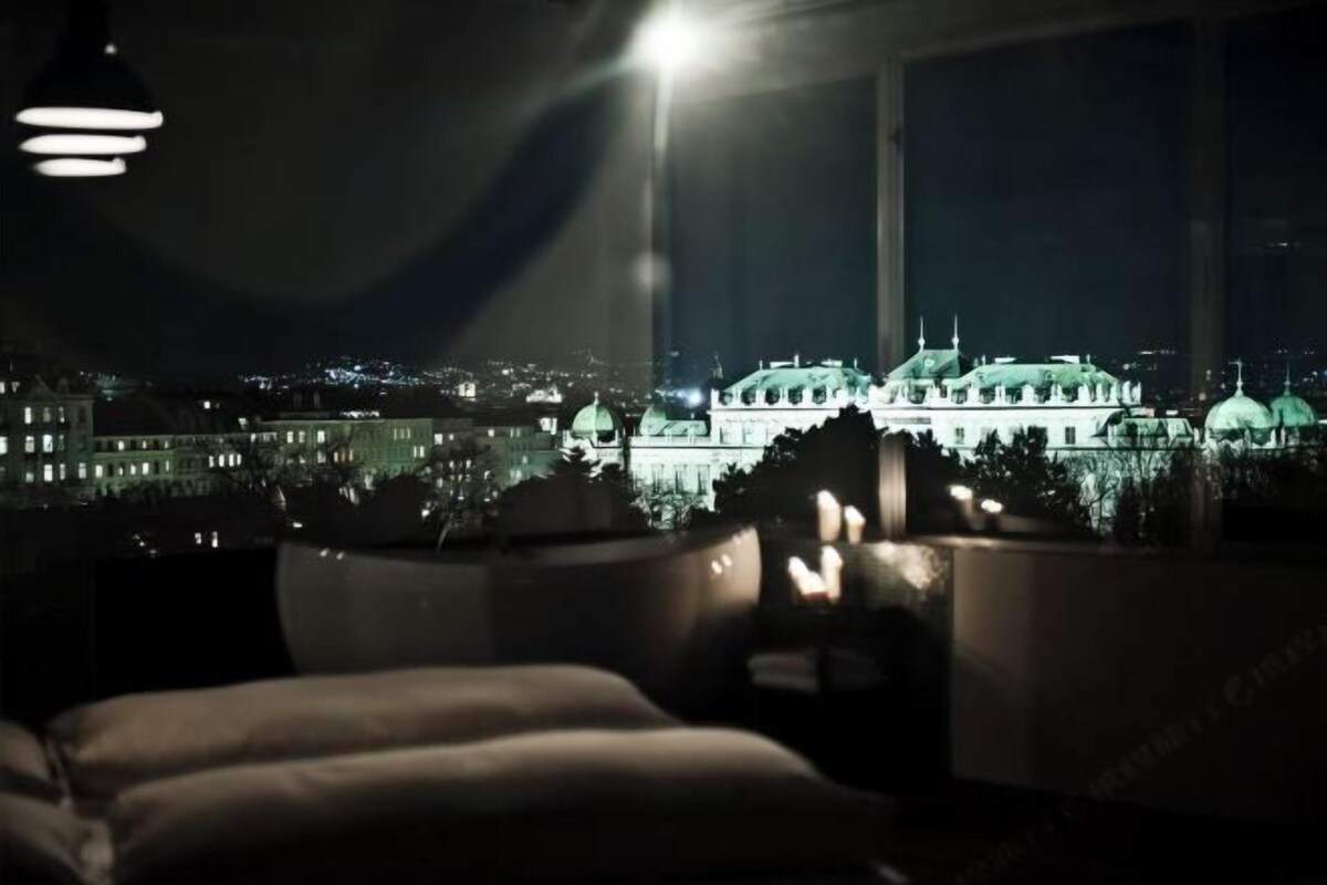 Une pièce dotée d’une grande fenêtre donnant sur une vue panoramique de la ville la nuit, avec, au premier plan, un bâtiment imposant surmonté de plusieurs dômes.
