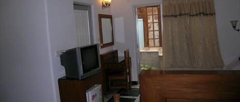 Una habitación con cama, televisión sobre un mueble bajo, un refrigerador pequeño, un espejo y una ventana con cortinas.