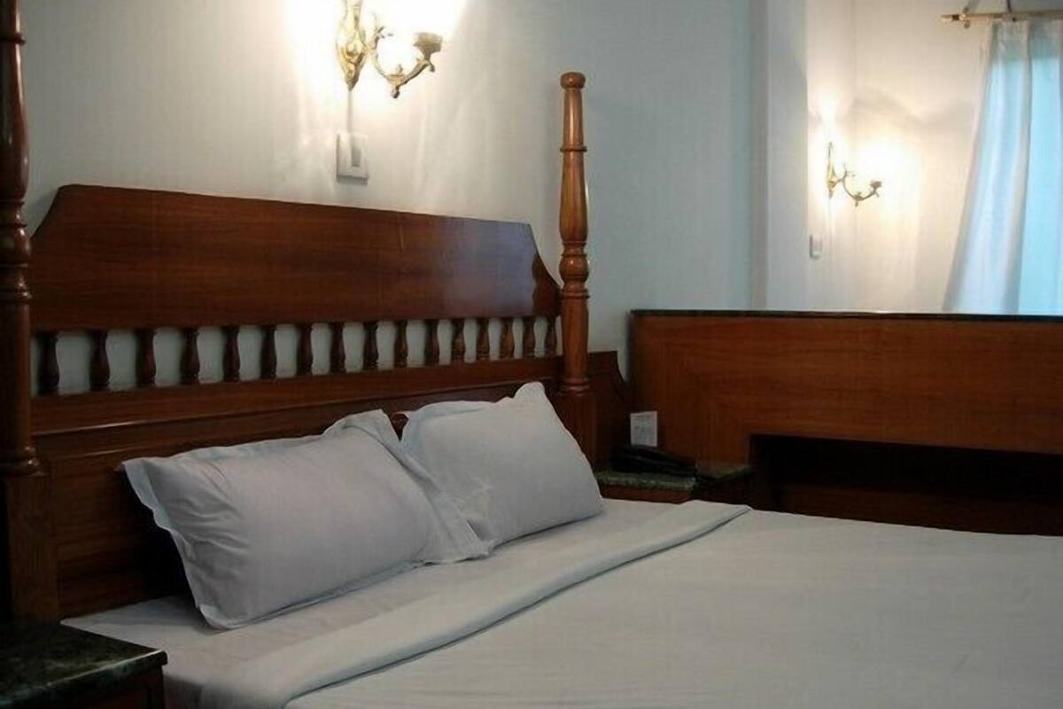 A wooden bed with a headboard and two pillows in a room with a nightstand and a wall-mounted light fixture.