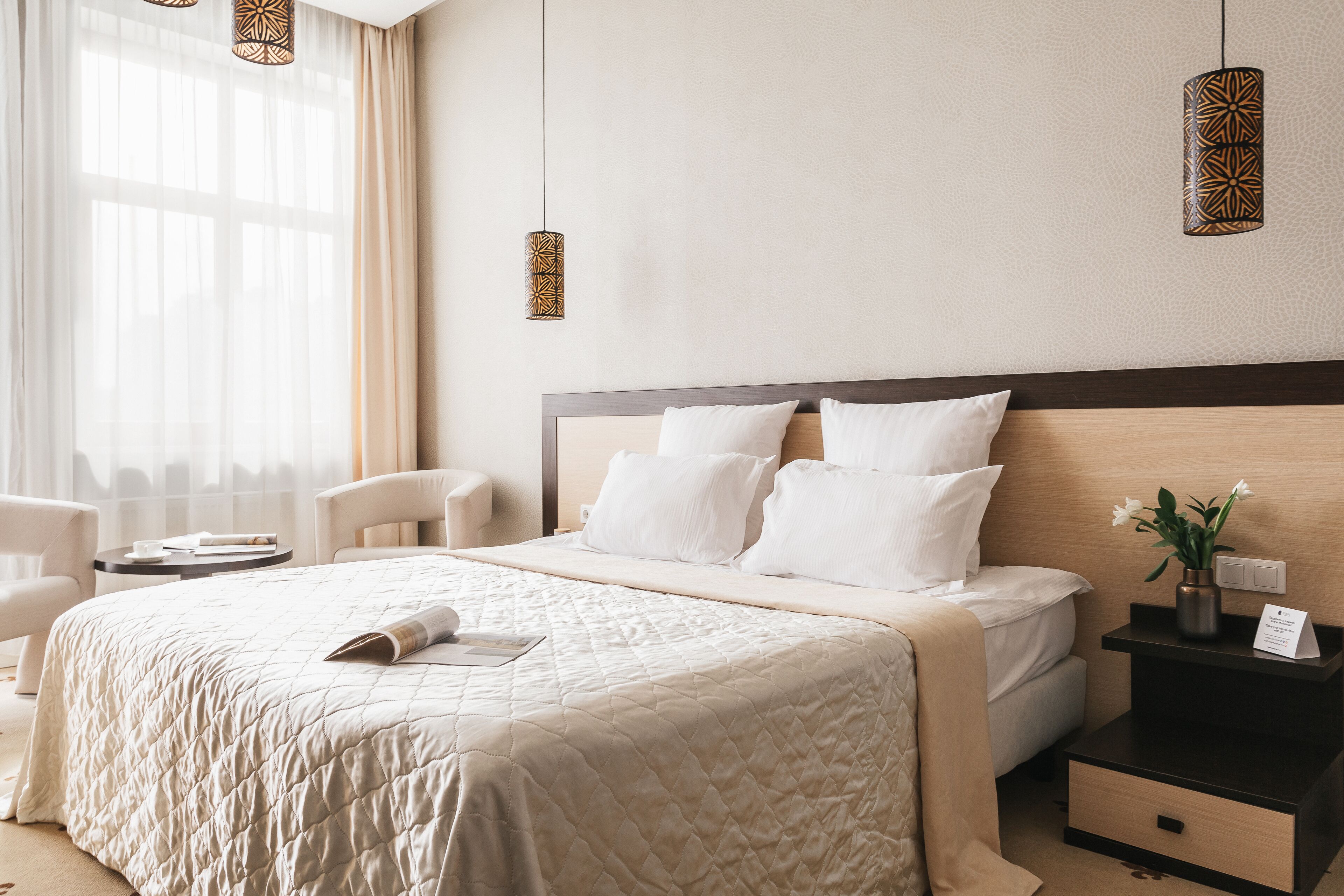 A hotel room with a large bed, a bedside table with a vase of flowers, and a window with sheer curtains.