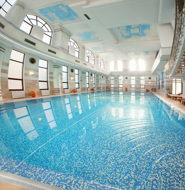 Una amplia piscina interior con agua cristalina y azul, rodeada de grandes ventanales y un techo alto con detalles decorativos.