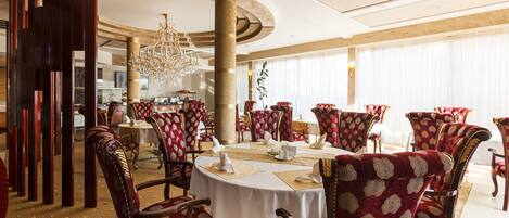 A formal dining area with round tables, red upholstered chairs, and a chandelier.