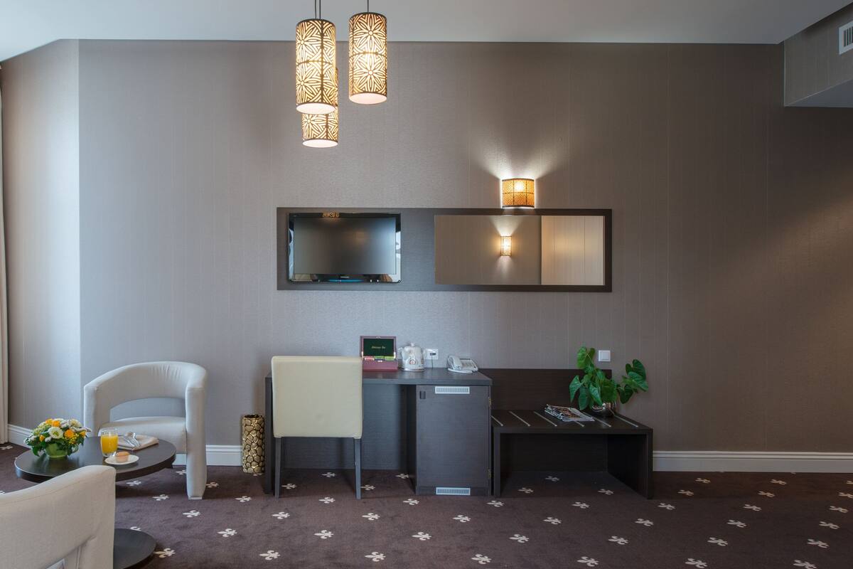 A hotel room with a seating area, a desk, a small refrigerator, a mirror, and a plant.