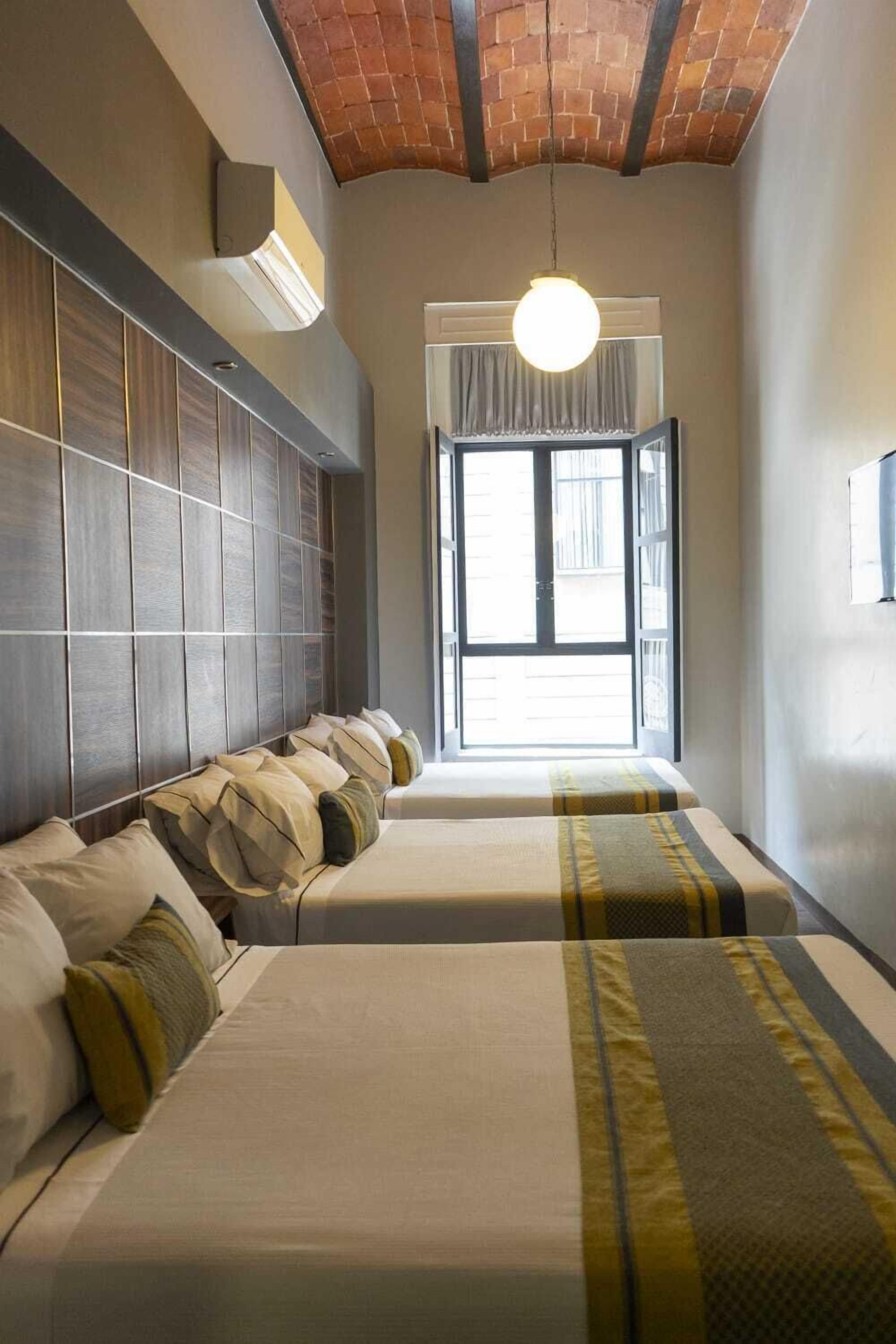 Une chambre d’hôtel avec deux lits, un plafond en briques, une grande fenêtre et un luminaire suspendu.