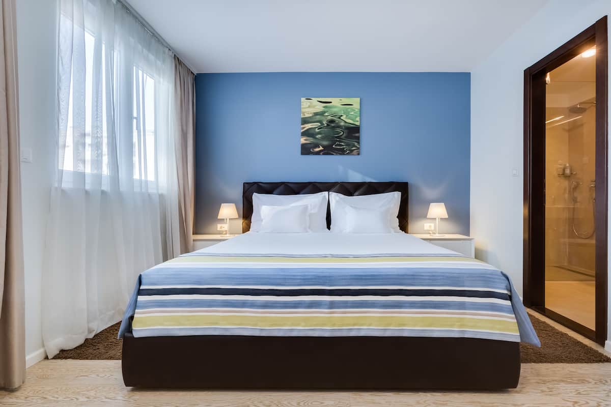 Quarto de hotel com uma cama grande, duas mesas de cabeceira com luminárias, uma janela com cortinas translúcidas e uma porta de vidro que dá para um espaço do chuveiro.