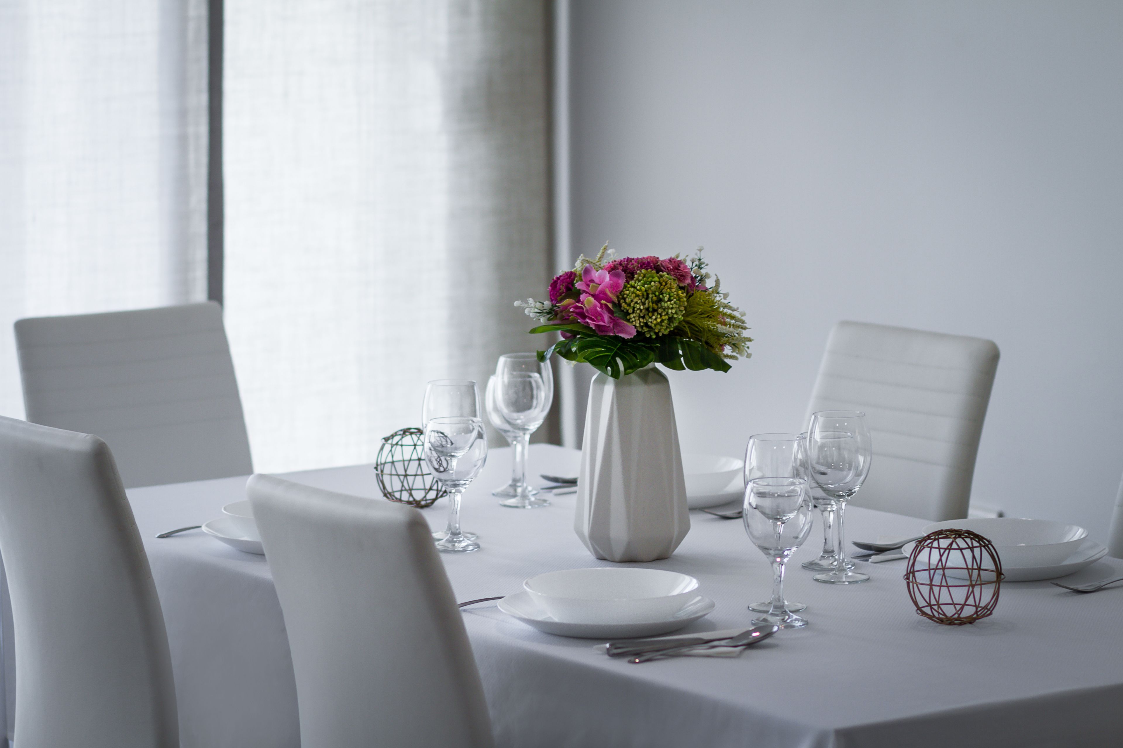 A dining table set with white plates, clear wine glasses, and a white vase with flowers.