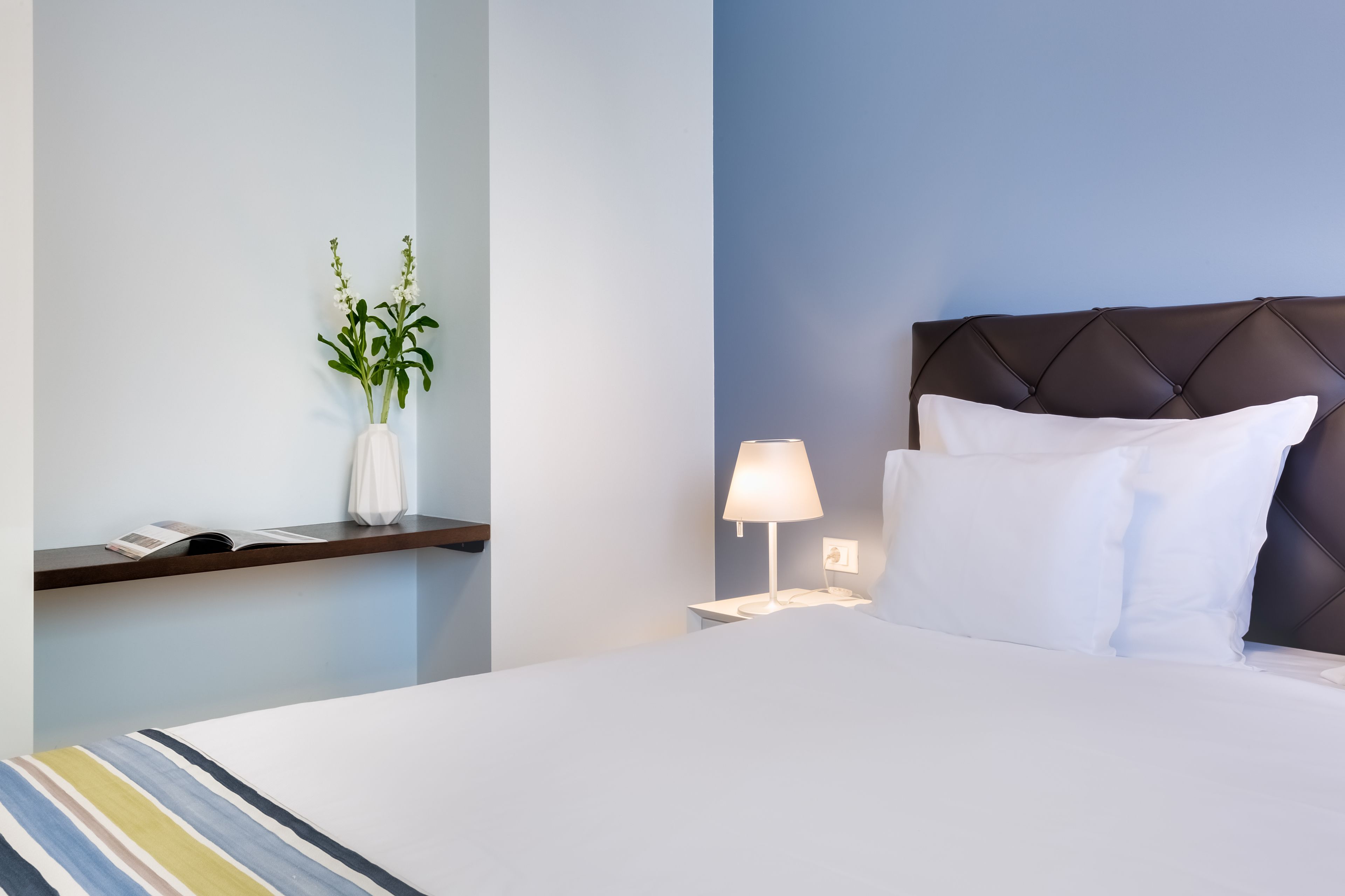 A hotel room with a bed, a nightstand, a lamp, a vase with flowers, and a shelf with a book and a phone.
