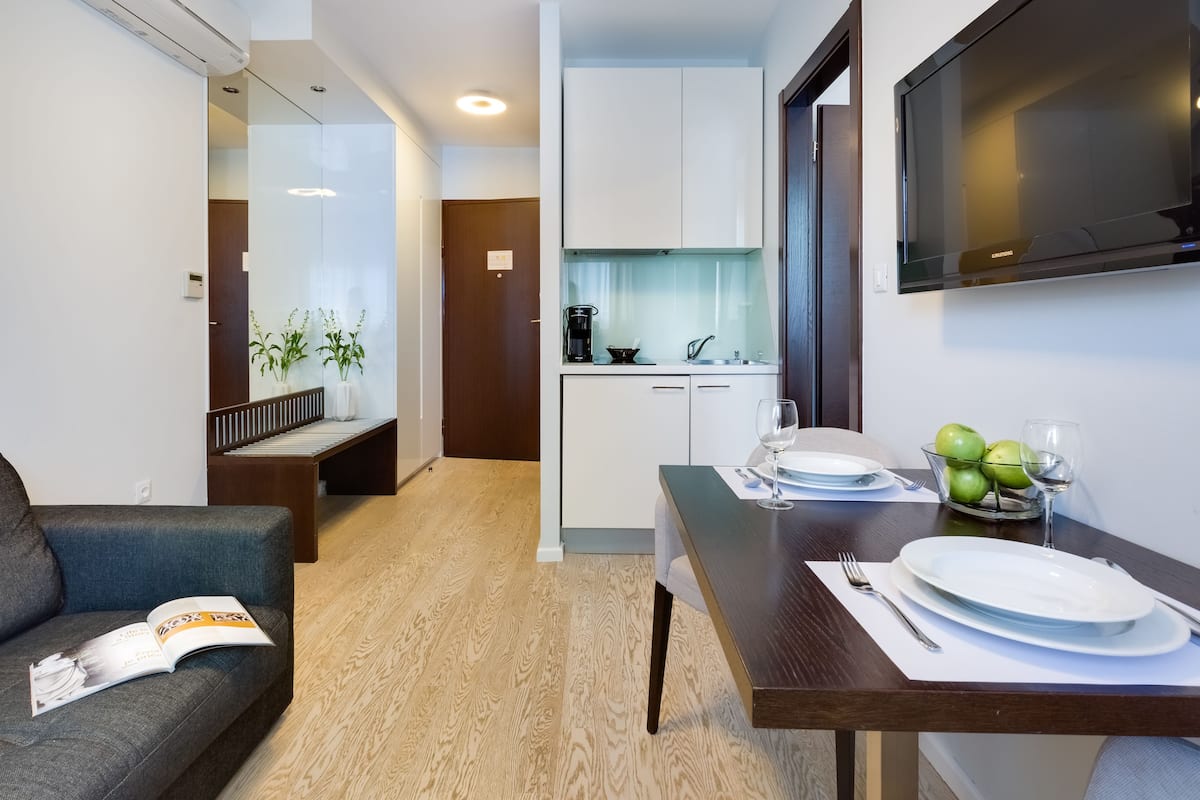 Uma sala de estar moderna com sofá, mesa de jantar posta com pratos e copos, área de cozinha com armários e uma TV de tela plana na parede.
