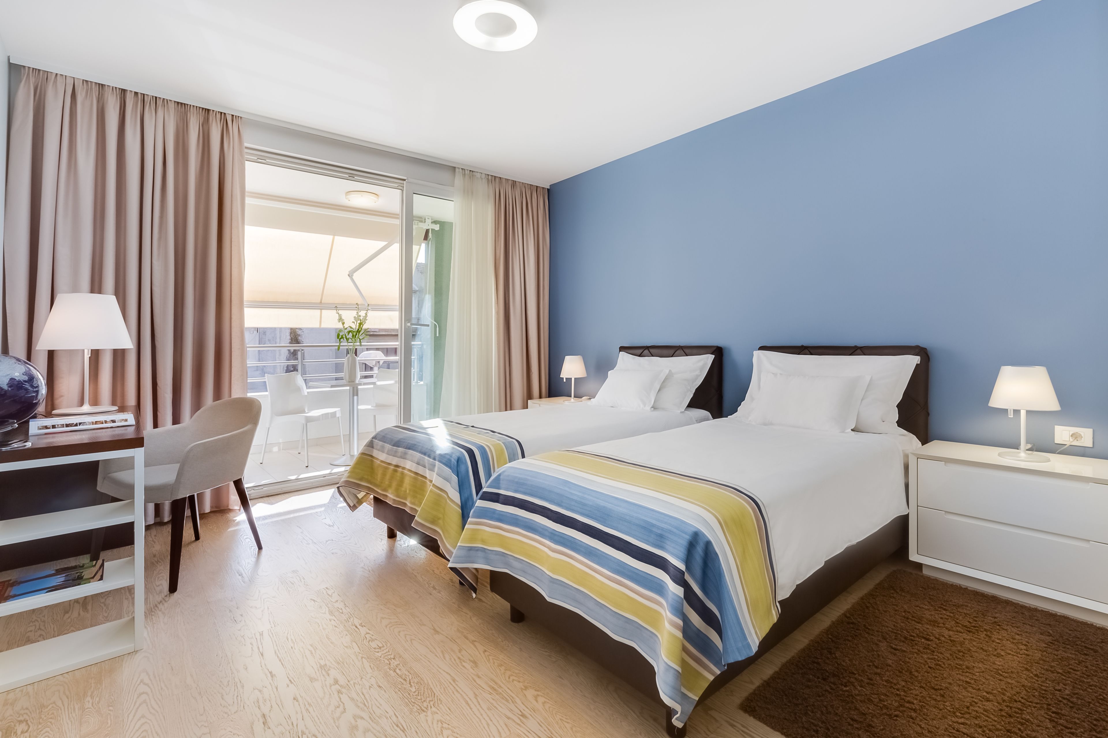 A hotel room with a bed, a desk with a chair, a laptop, and a window with curtains.