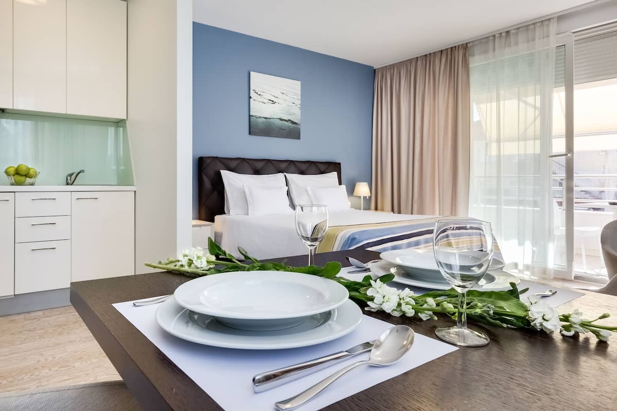 Quarto de hotel com uma mesa de jantar bem posta, cama com roupa de cama branca, vista da cidade e cozinha moderna com armários brancos.