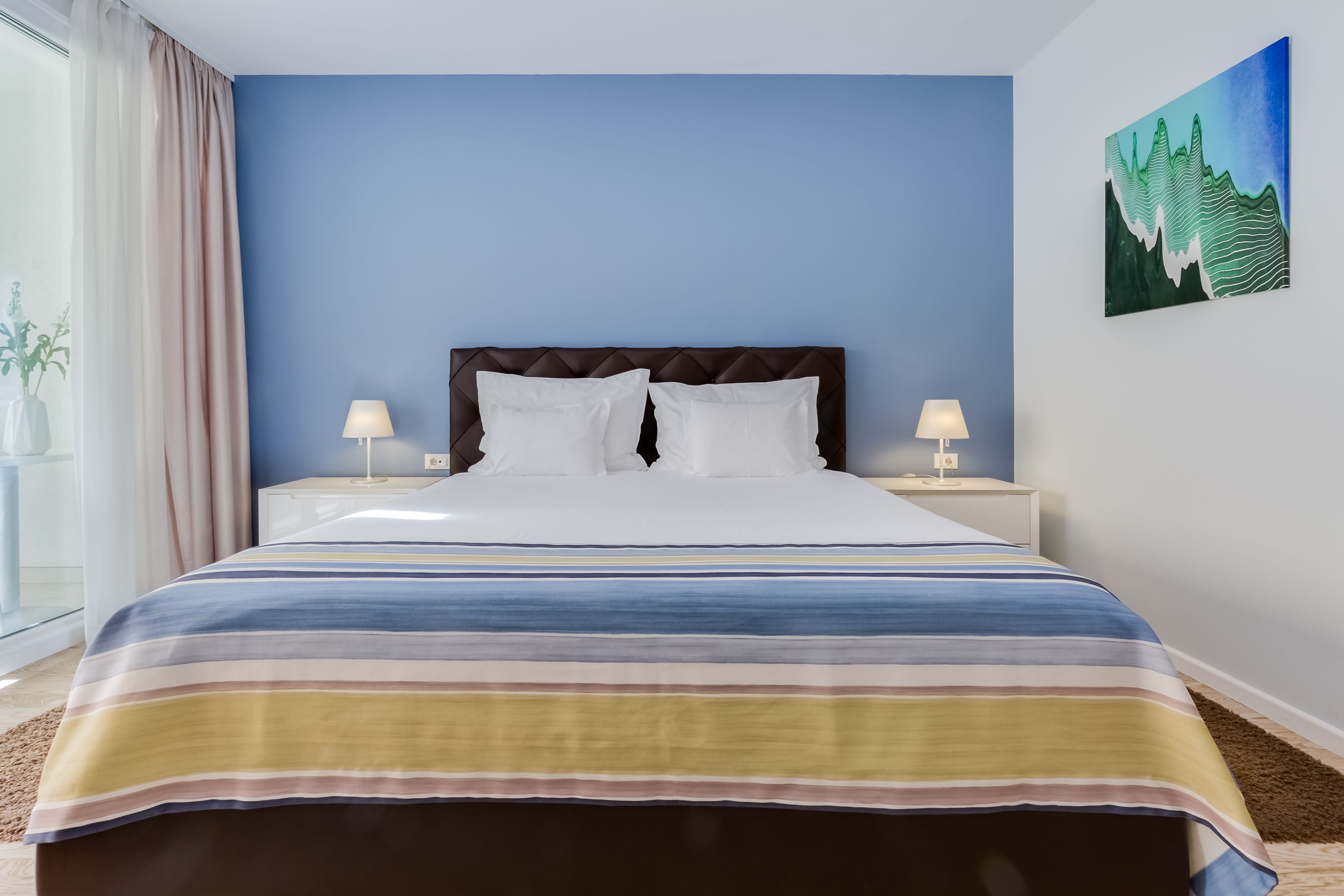 Ein Hotelzimmer mit Bett, zwei Nachttischlampen, einem Bild an der Wand und einem Fenster mit Vorhängen.