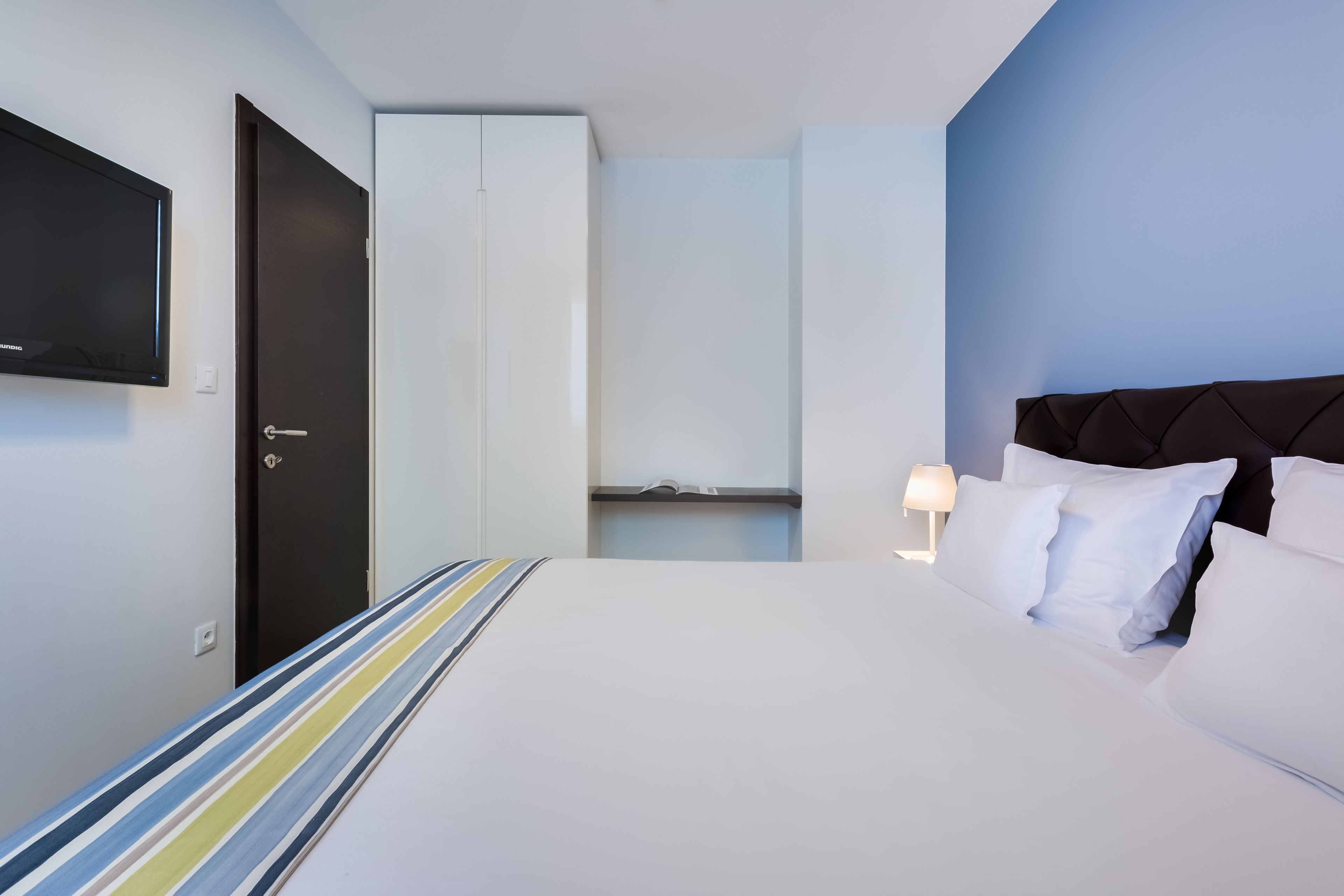 A hotel room with a bed, a TV mounted on the wall, a wardrobe, and a bedside table with a lamp.