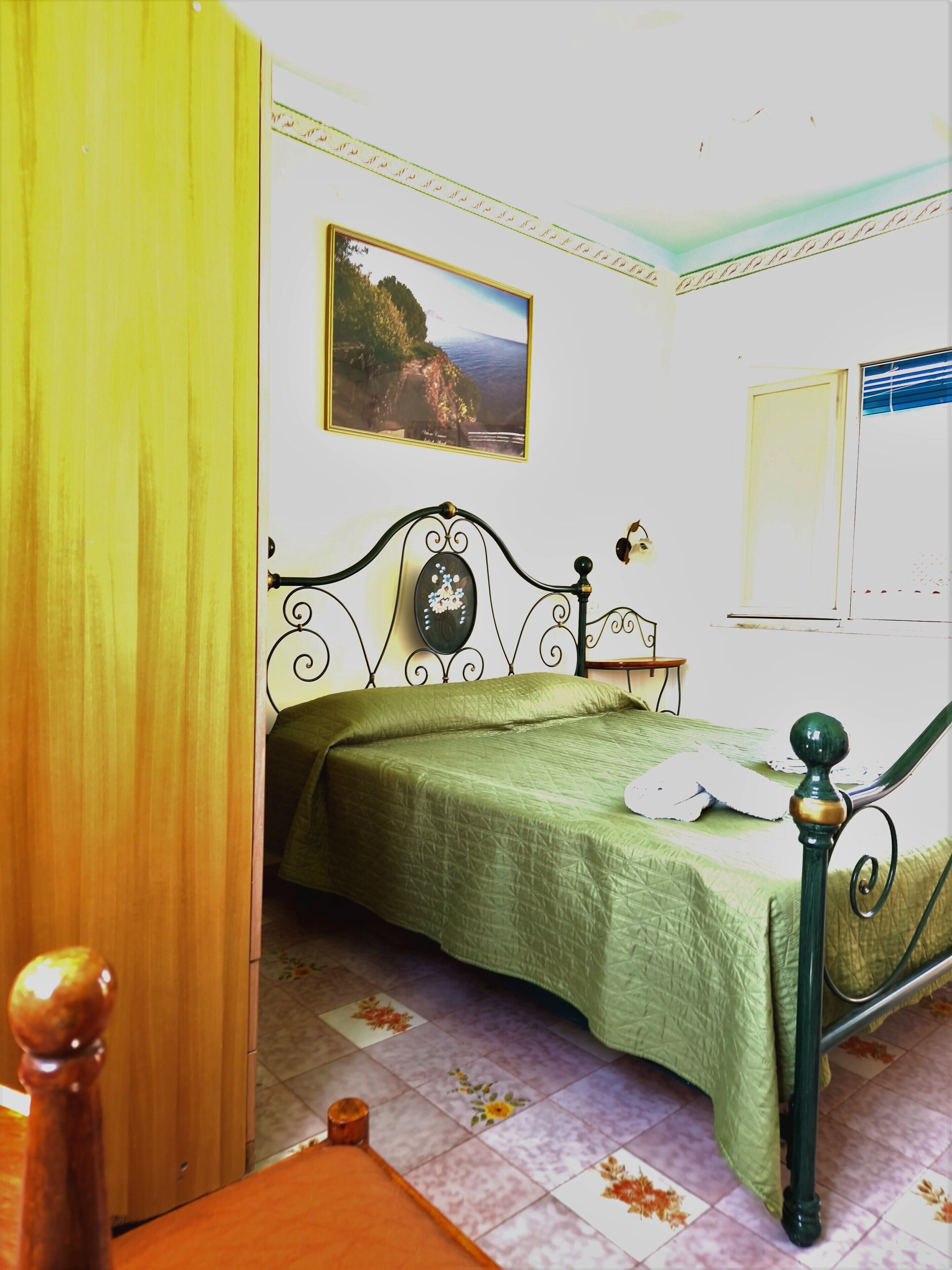 A bedroom with a bed, a painting on the wall, and a window with blinds.