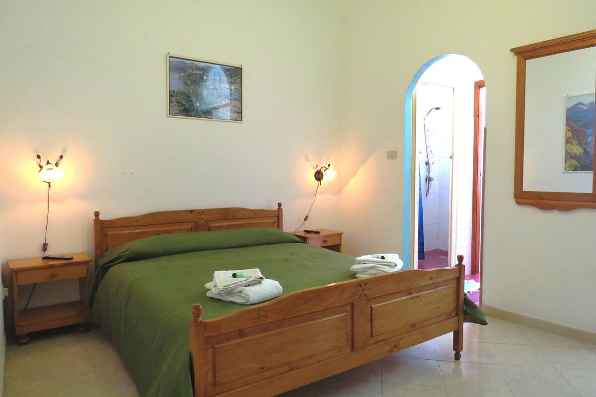 A bedroom with a wooden bed, green bedspread, two bedside lamps, a painting on the wall, and a bathroom visible through an archway.