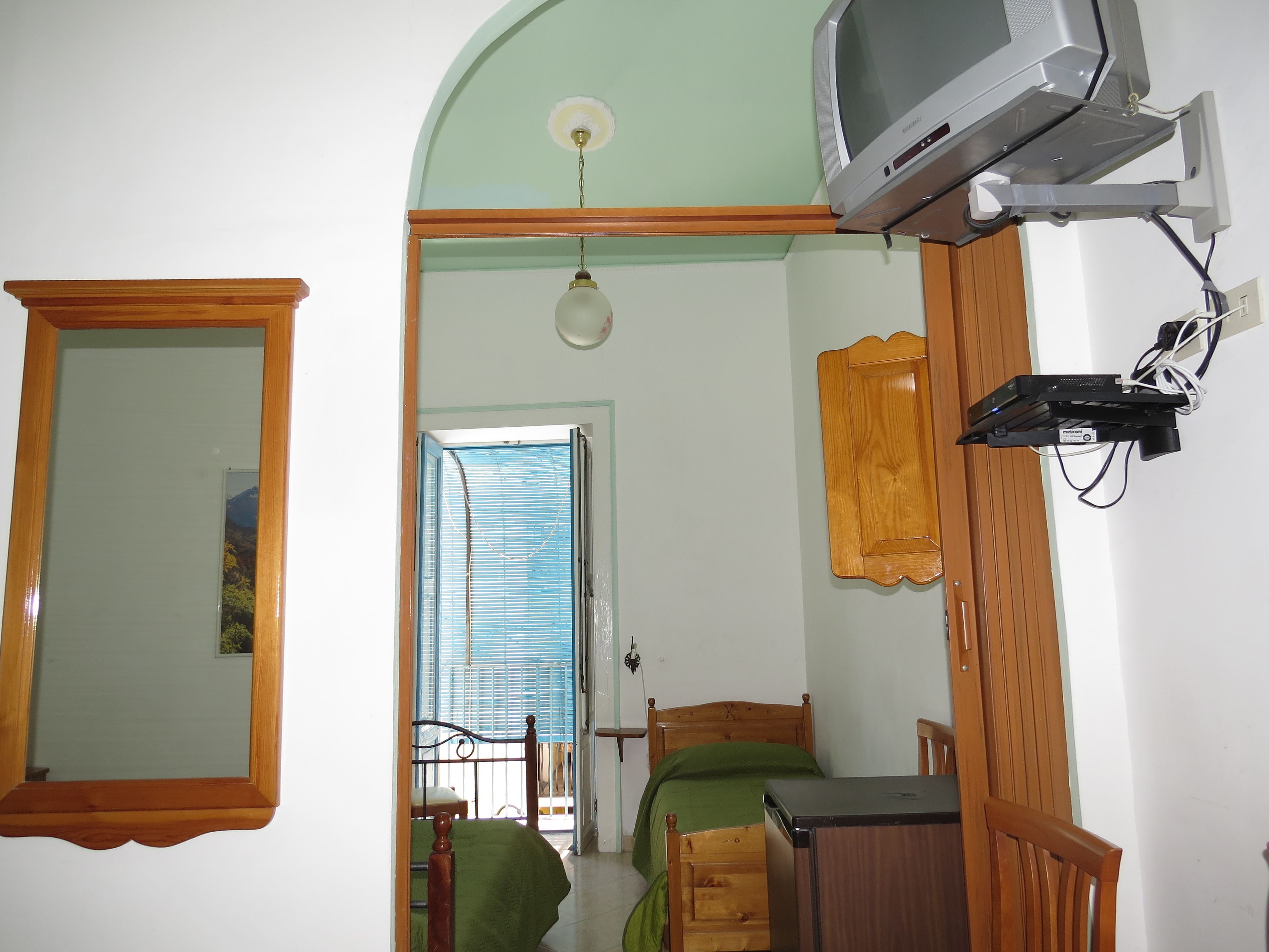Un dormitorio con una cama, una silla de madera, un escritorio, un televisor fijado a la pared y una ventana con persianas.