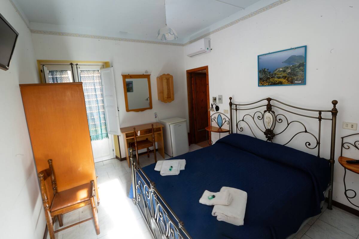A bedroom with a bed, a desk, a chair, a television, and a painting on the wall.