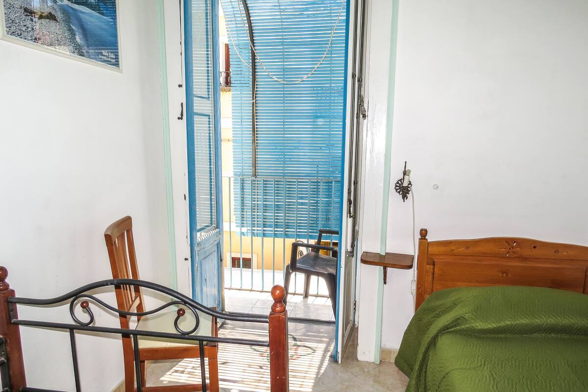 A room with a bed, a chair, a table, and a balcony with blue shutters.