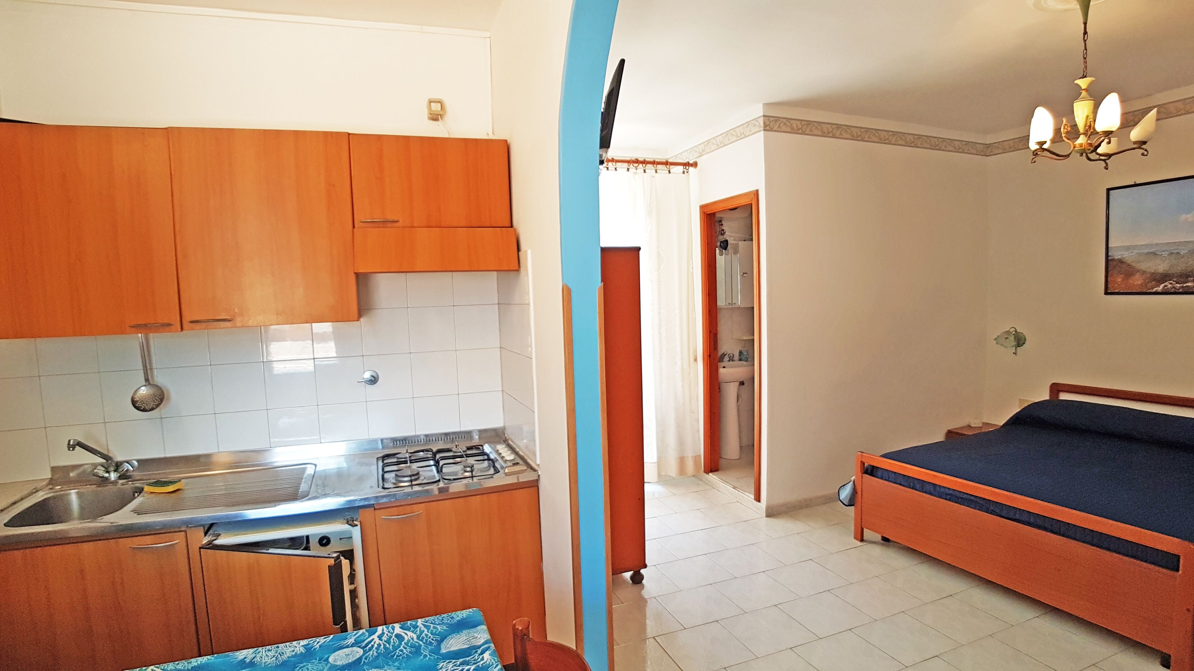 A compact kitchen with wooden cabinets, a sink, and a stove. There is a bed with a blue mattress and a wooden headboard in the adjacent room.