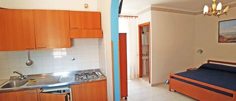 A compact kitchen with wooden cabinets, a sink, and a stove. There is a bed with a blue mattress and a wooden headboard in the adjacent room.