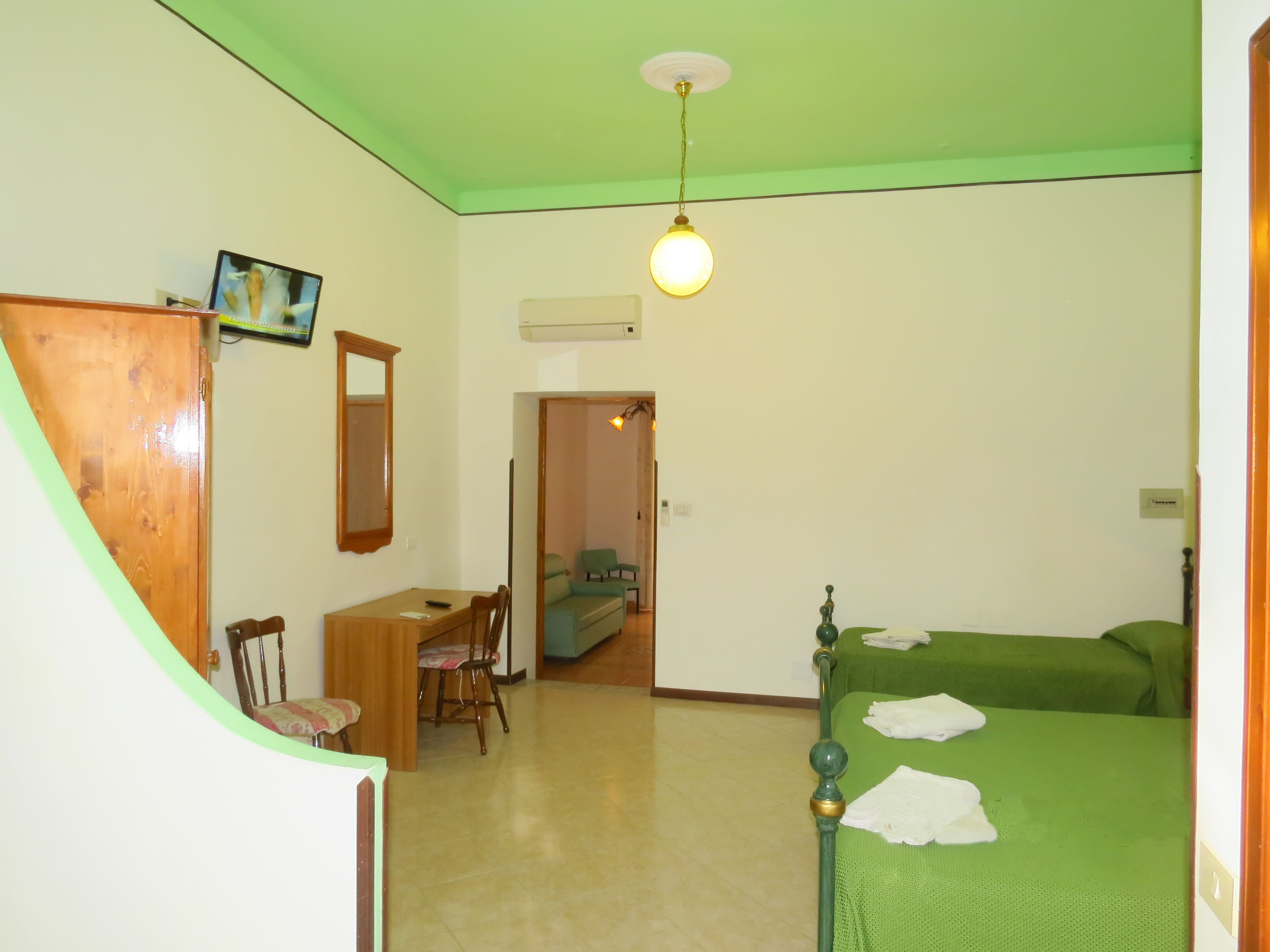 A room with a bed, a desk, and a chair. There is a mirror on the wall and a light fixture above the bed.