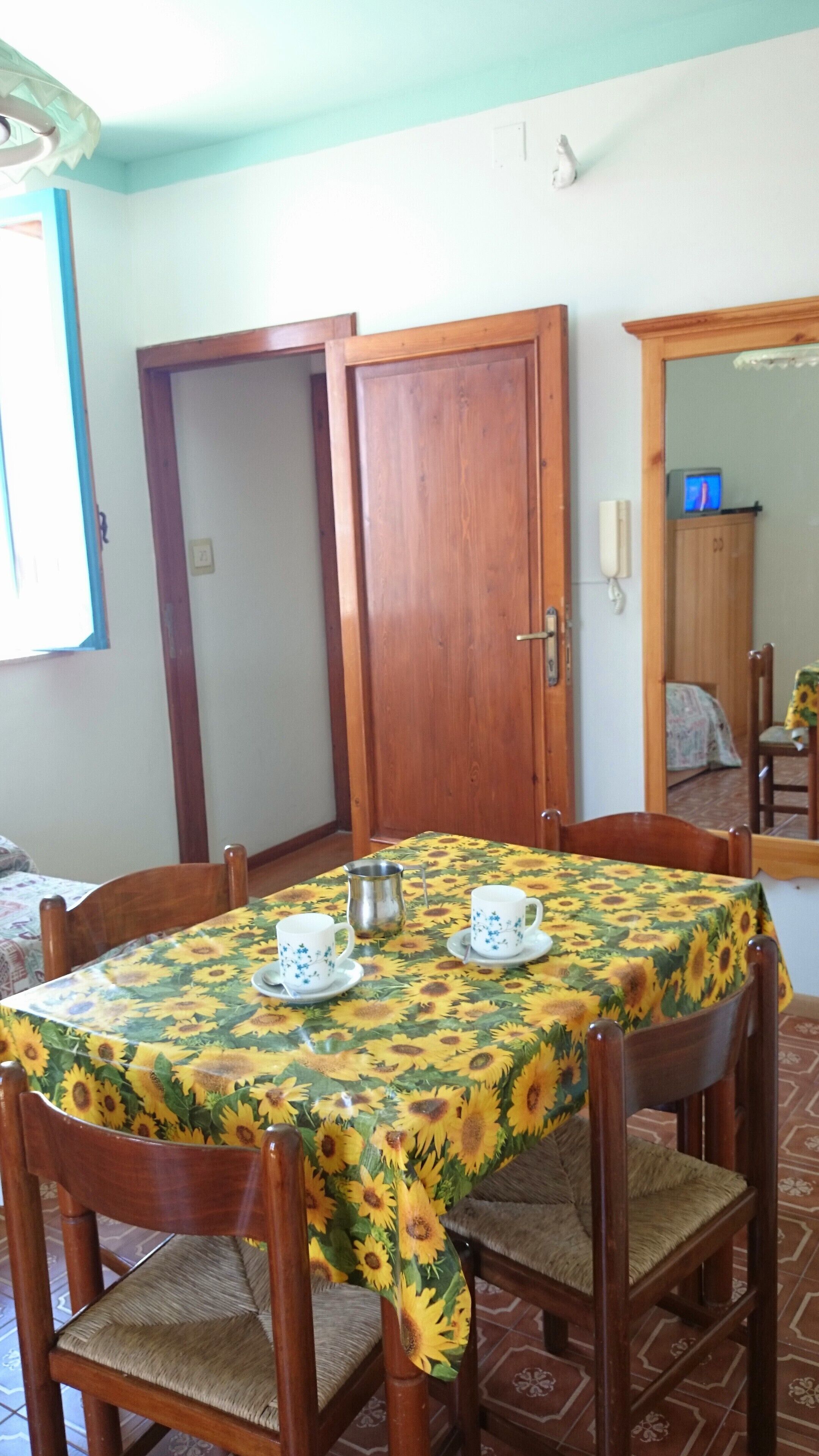 A dining area with a table covered in a sunflower tablecloth, surrounded by wooden chairs with cushioned seats.