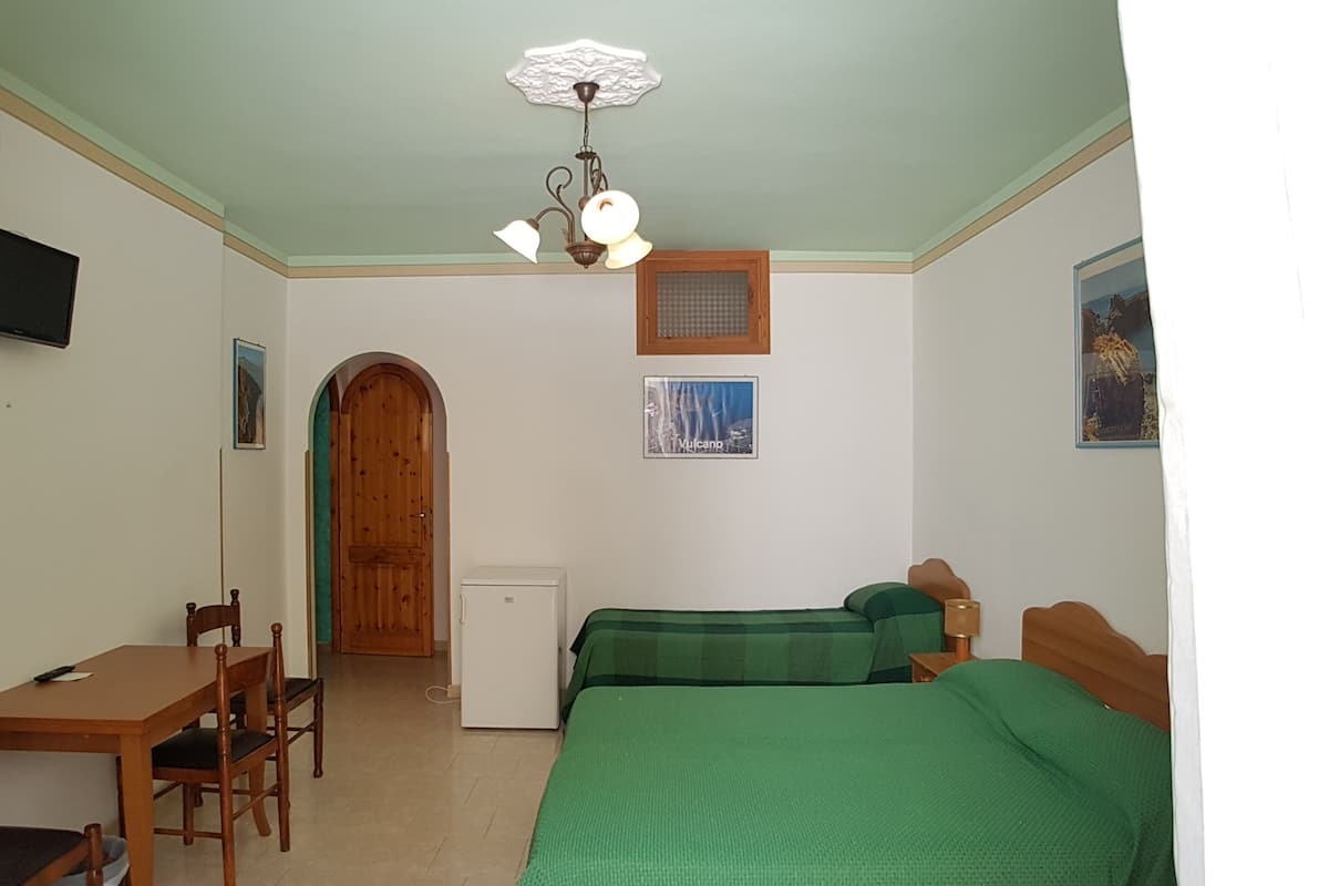 A room with a wooden door, a small table with chairs, a bed with green bedding, a ceiling fan, and a wall-mounted TV.