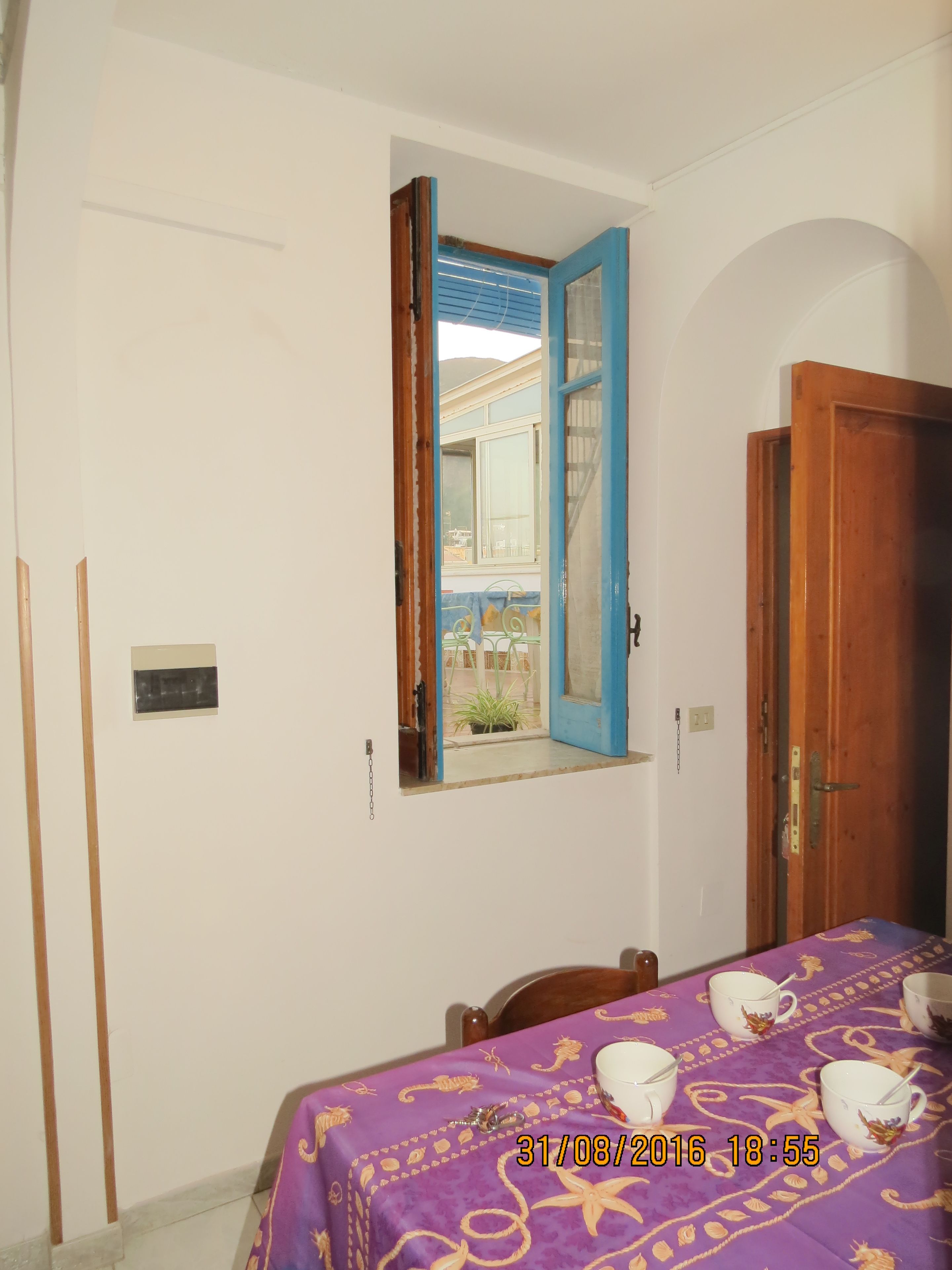 A room with a bed, a table with cups, a window with an open door, and a view of a building outside.