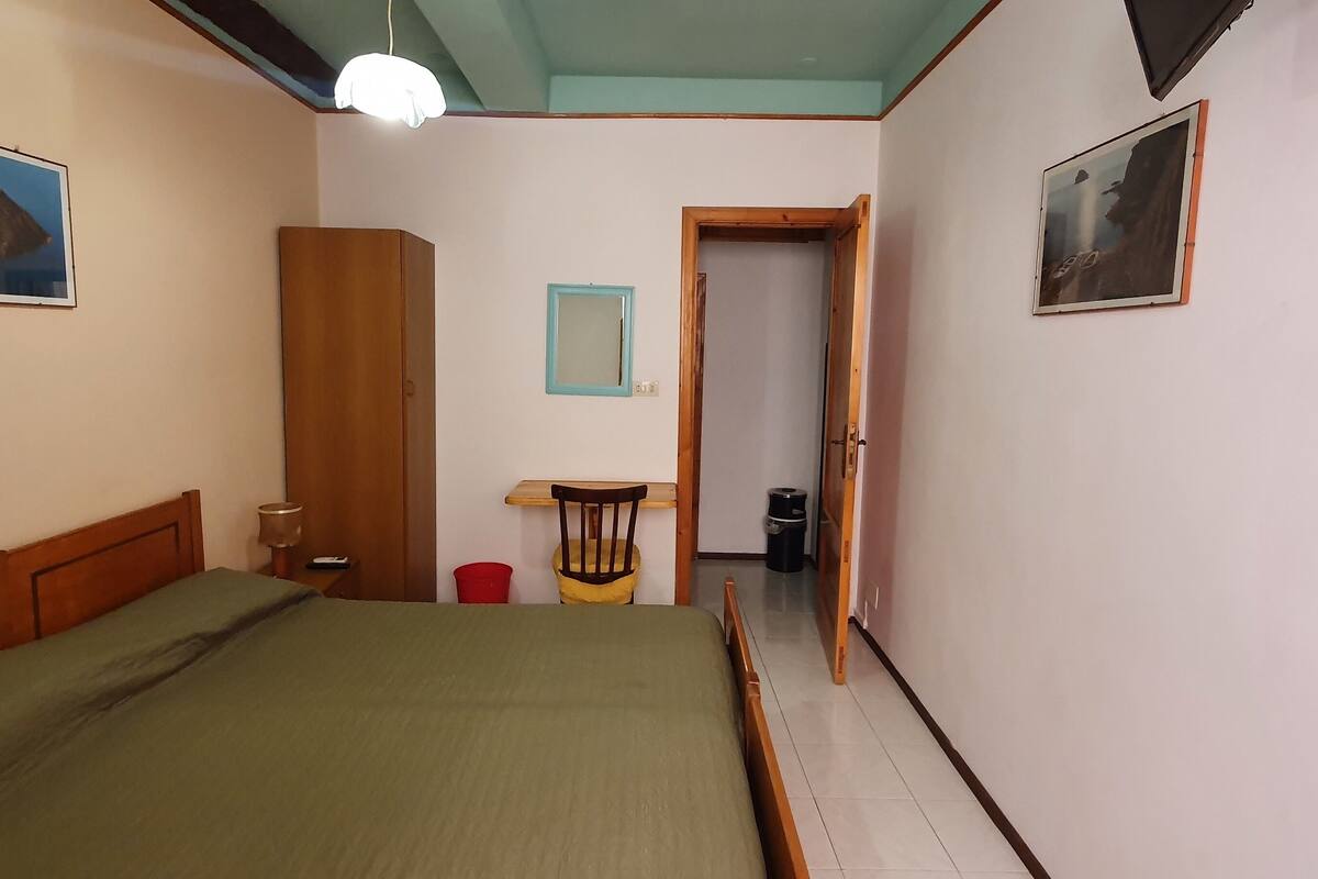 A bedroom with a bed, a wooden wardrobe, a small table, a chair, a red trash bin, and a door leading to another room.