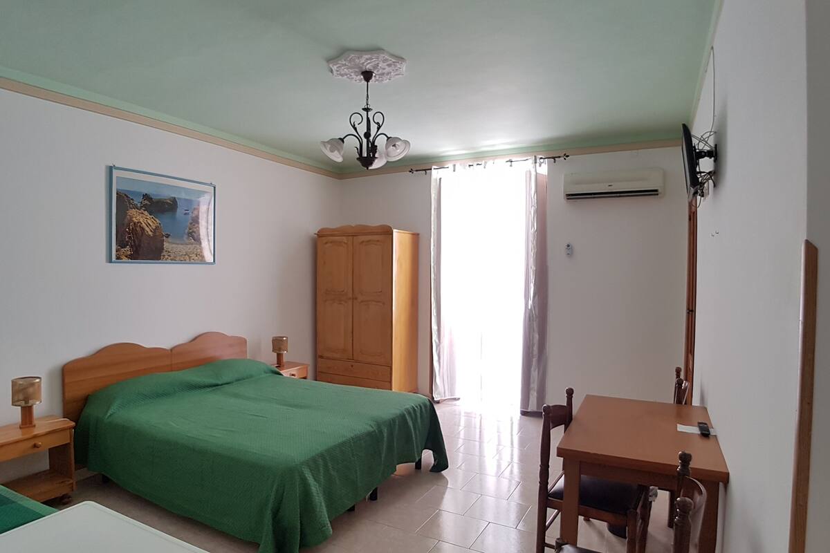A bedroom with a bed, a wooden wardrobe, a desk with a chair, a nightstand, a lamp, and a ceiling light fixture.