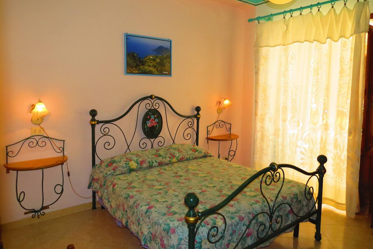 A bedroom with a bed, two bedside tables, a wall-mounted lamp, a picture, and a window with curtains.