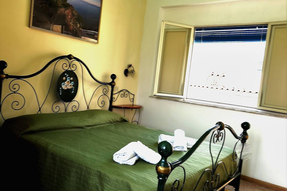 A bedroom with a green bedspread, a metal-framed bed, a painting on the wall, a window with blinds, and a ceiling light.