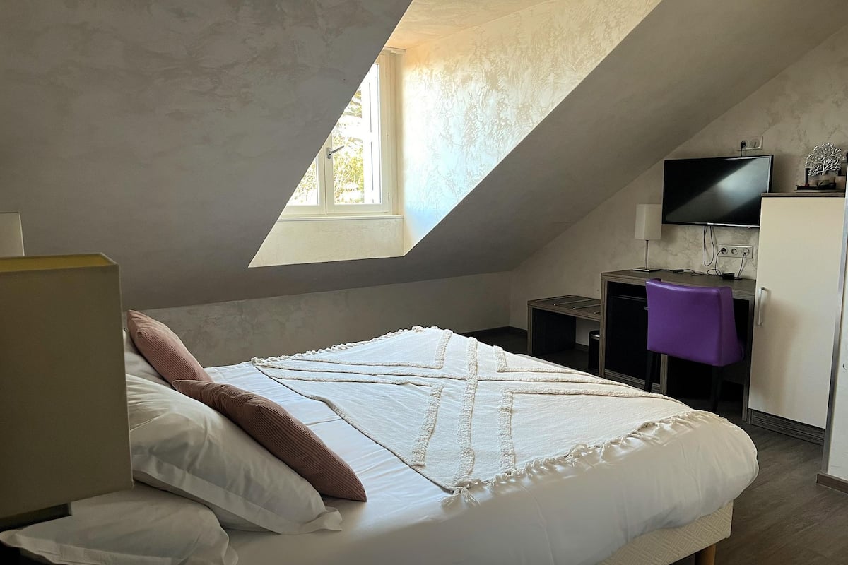 Een slaapkamer met een schuine kap, een bed met witte beddengoed, een bureau met een computer, een televisie en een paarse stoel.