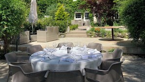 Un espace de restauration en plein air, avec des chaises et des tables en osier disposées pour un repas, entouré d’une végétation luxuriante et avec une maison située sur une colline en arrière-plan.