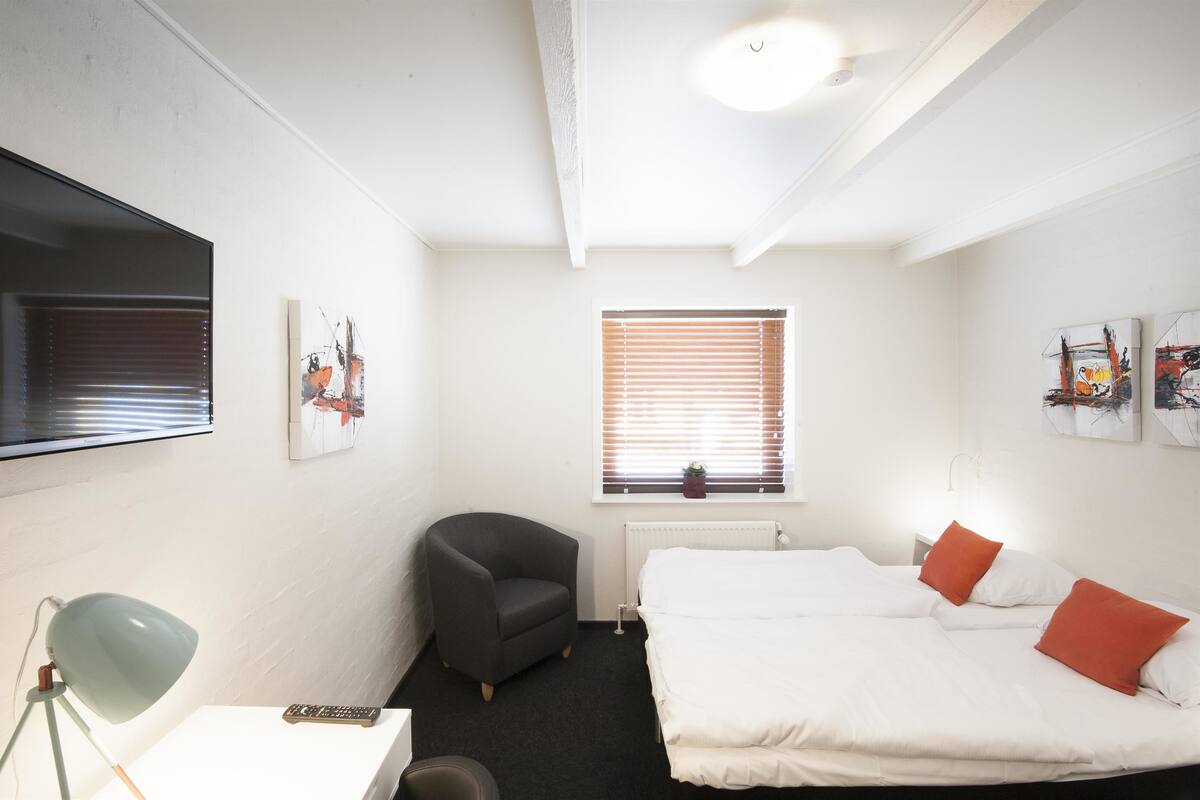 Quarto de hotel com cama, uma cadeira, escrivaninha com abajur, televisão e obras de arte nas paredes.