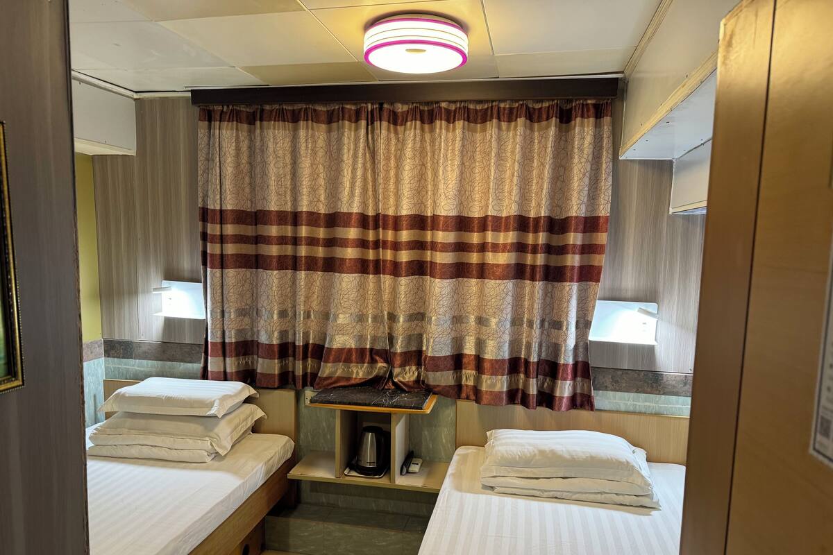 A small, compact hotel room with two single beds, a small table with a kettle, and a ceiling light fixture.
