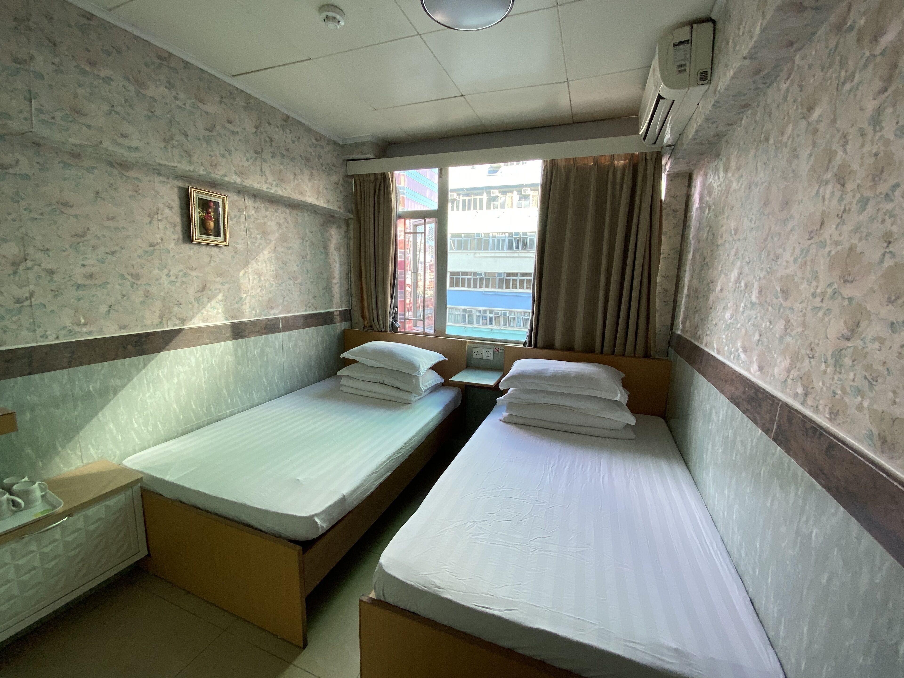 Habitación de hotel con dos camas individuales, una mesita de noche pequeña, un calentador fijado a la pared y una ventana con cortinas.