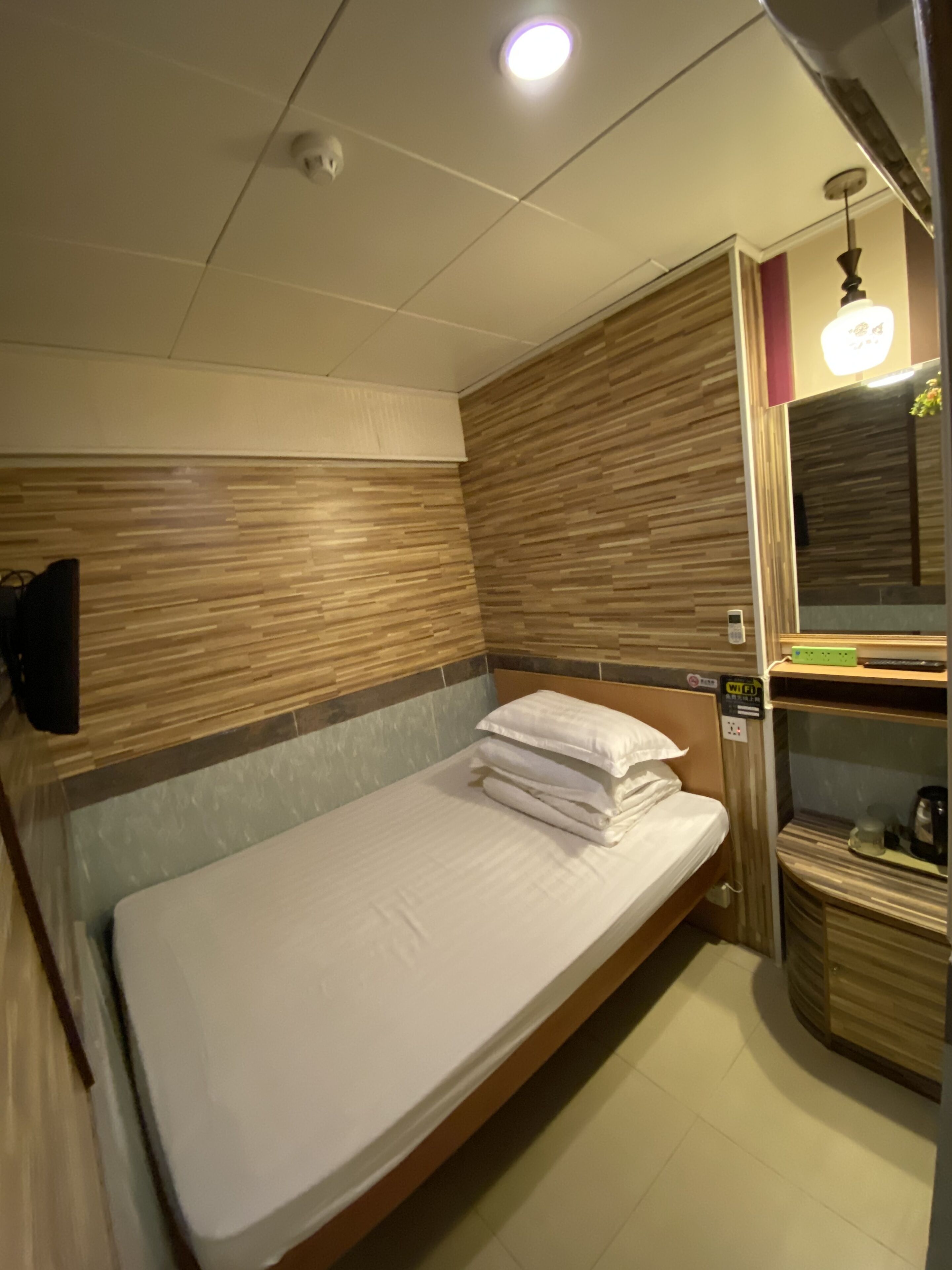 A single bed with a wooden headboard, a bedside table with a lamp, a mirror, and a small shelf with a kettle.