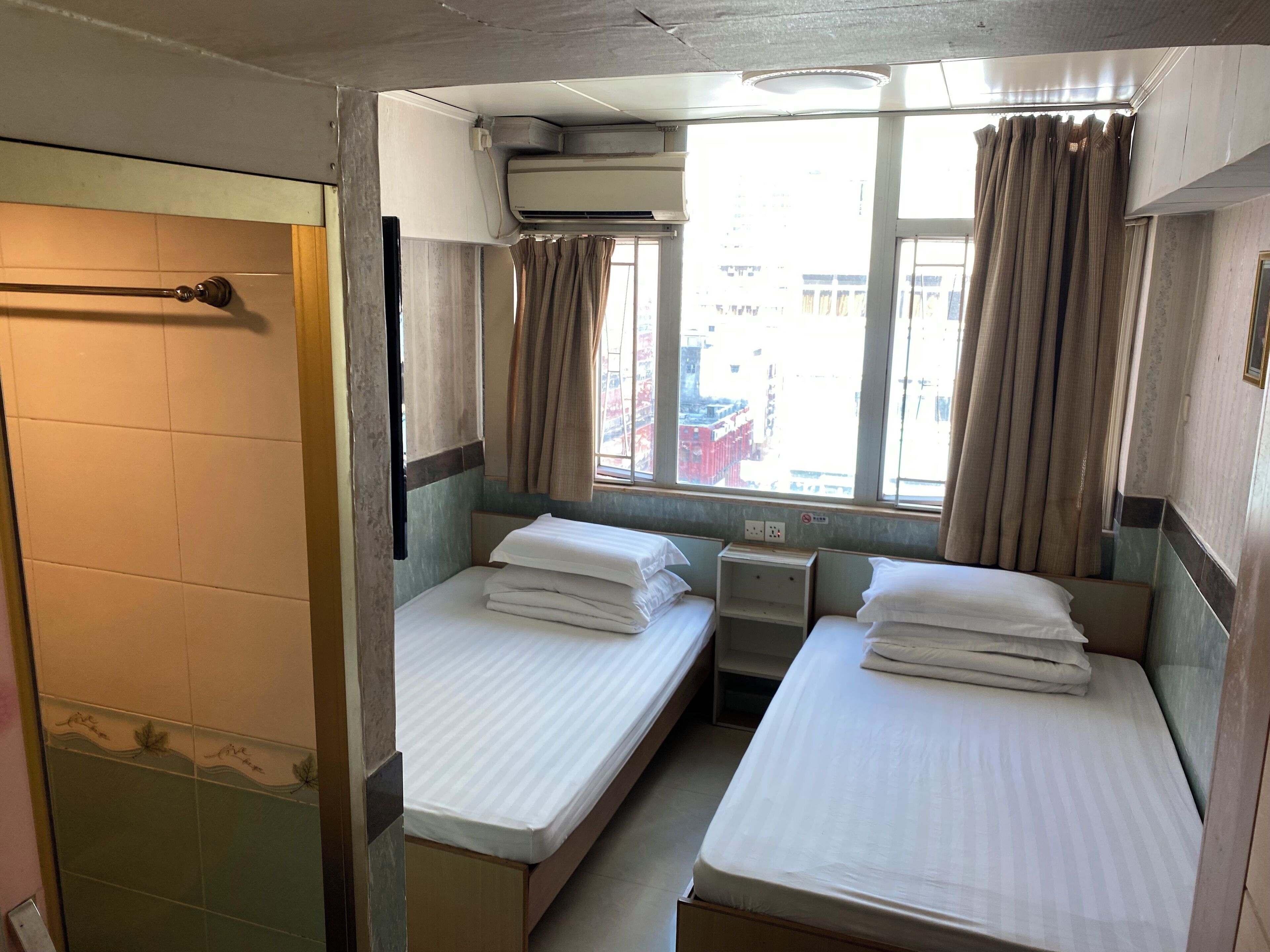 Ein kleines, kompaktes Hotelzimmer mit zwei Einzelbetten, einem Fenster mit Vorhängen, einer Klimaanlage und einem kleinen Nachttisch.