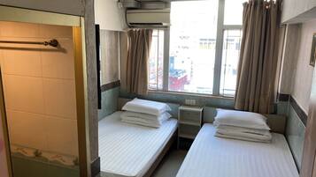 A small, compact hotel room with two single beds, a window with curtains, an air conditioning unit, and a small bedside table.