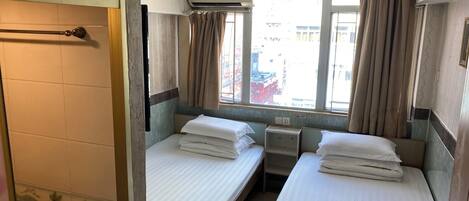 Habitación de hotel pequeña y funcional con dos camas individuales, una ventana con cortinas, un equipo de aire acondicionado y una mesita de noche.