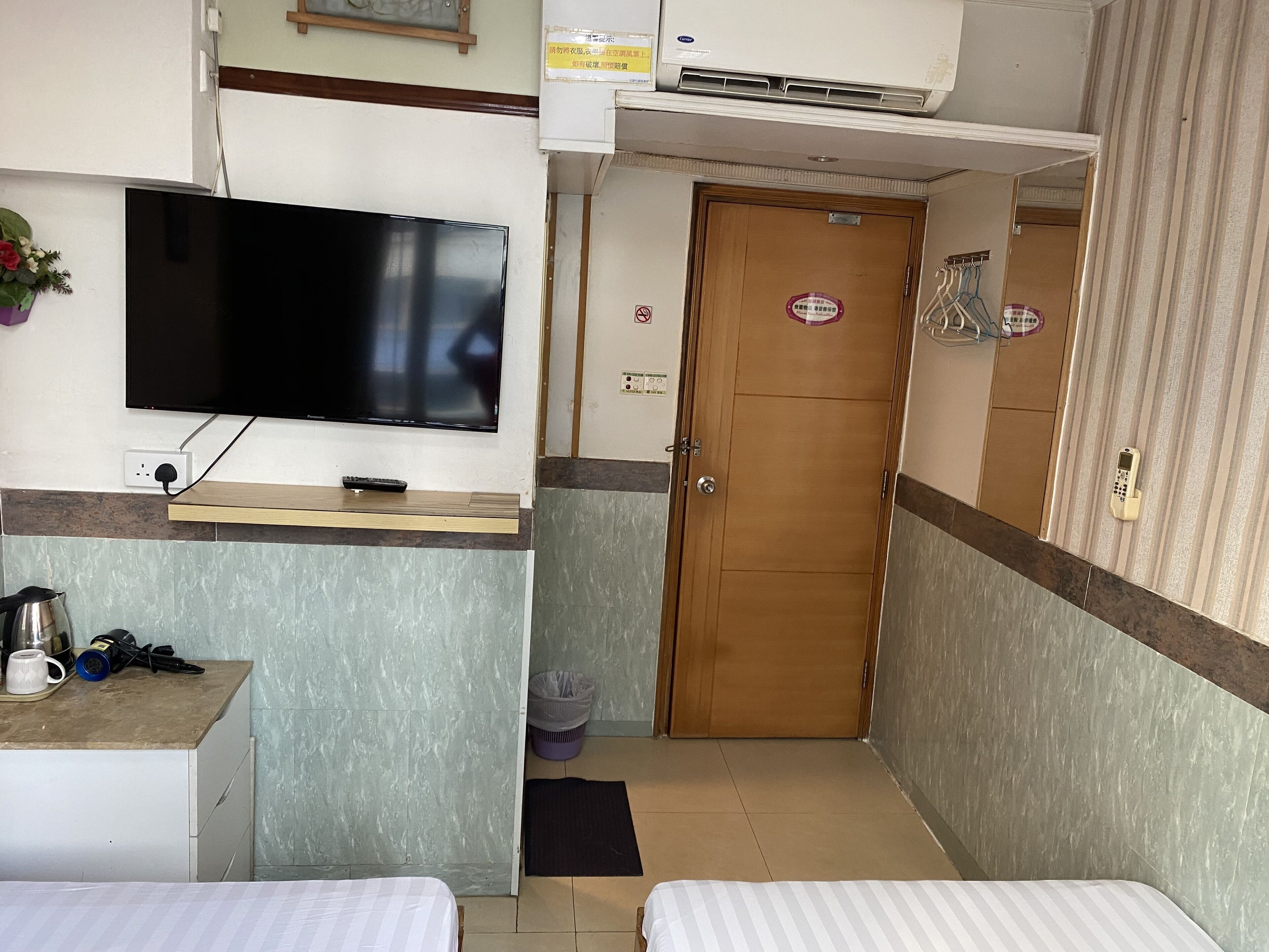 A hotel room with a flat-screen TV mounted on the wall, a wooden door, a small desk with a remote control, and two beds.