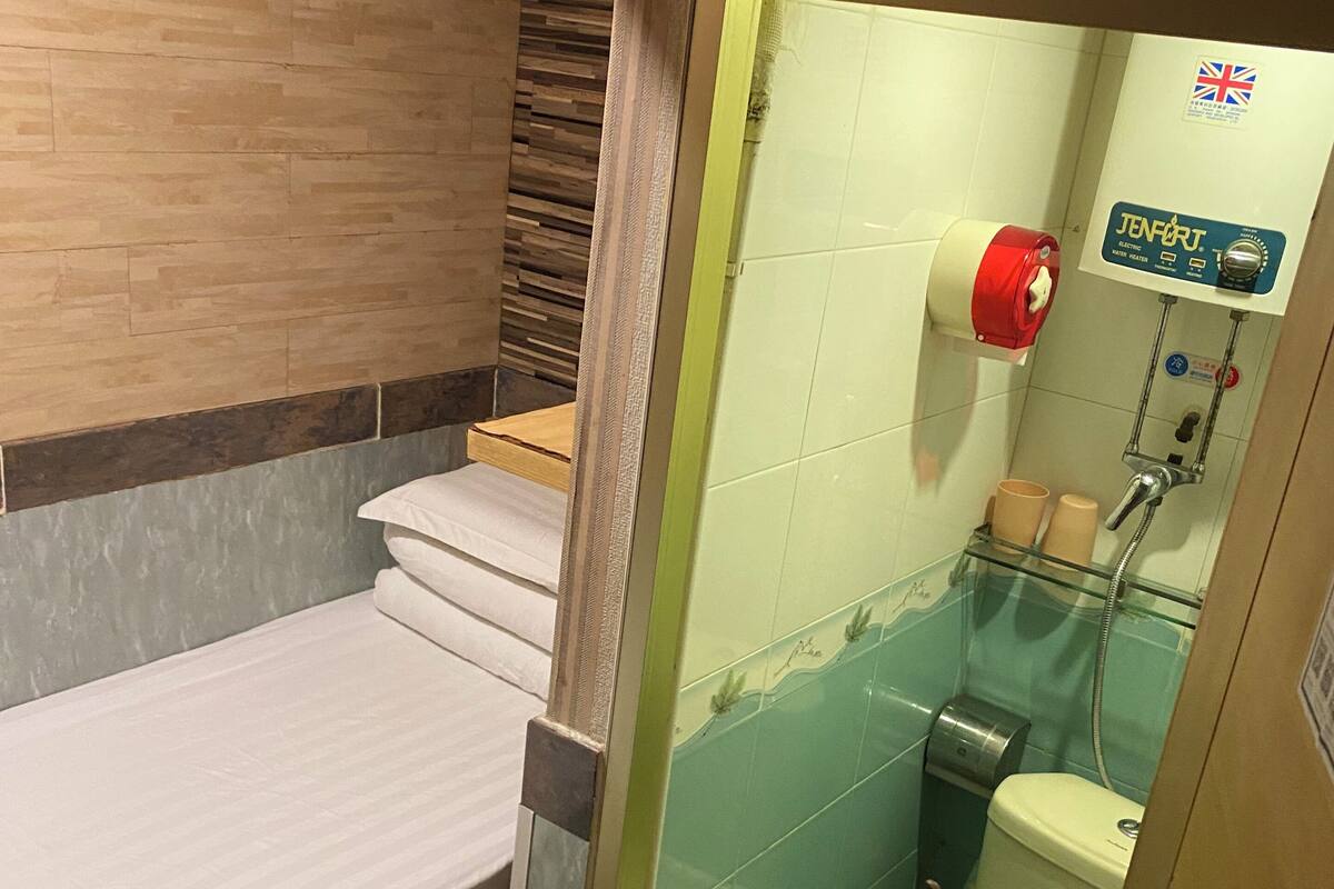 A small, compact bathroom with a toilet, a shower, and a sink. There is a bed with a white mattress and a wooden headboard.